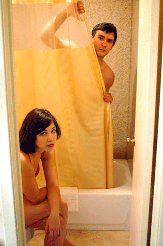 man peeking from behind shower curtain, woman sitting nearby