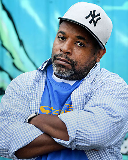 a middle-aged black man, arms crossed, in a ballcap, standing against a colorful wall